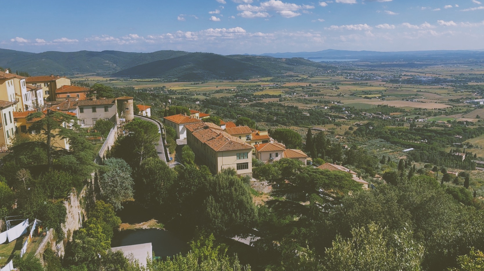 Italian countryside with white buildings with orange tiled roofs in the foreground and lush hills in the background.